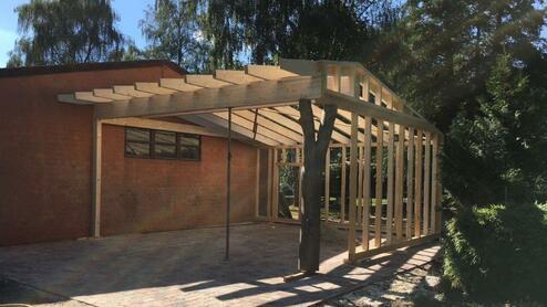Aufbau eines Holz-Carports neben einem Gebäude Konstruktion eines Carports aus Holz mit einem teilweise fertiggestellten Dach, nahe an einem gemauerten Gebäude.